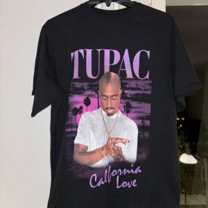 Svart T-shirt med Tupac-tryck - Säljer en svart T-shirt med ett stort tryck av Tupac och texten 'California Love' i lila och rosa. Perfekt för alla som älskar klassisk hiphop och Tupac.