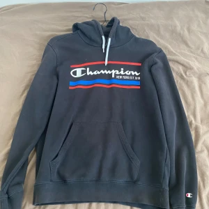 Svart hoodie från Champion - Säljer en svart hoodie från Champion med deras ikoniska logga i vitt, rött och blått på bröstet. Den har en känguruficka och justerbar huva med vita snören. Perfekt för en avslappnad stil eller träning.