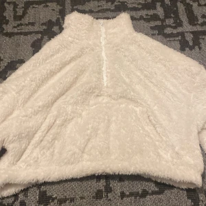 Vit fluffig tröja med dragkedja - Supermysig vit fluffig tröja som är perfekt för kalla dagar. Den har en halv dragkedja framtill och en stor ficka för att värma händerna. Tröjan är långärmad och gjord av ett mjukt, pälsliknande material som känns som att ha på sig ett moln. Perfekt för höst och vinter!