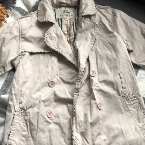 Beige trenchcoat från Burberry - Säljer en klassisk beige trenchcoat från Burberry i storlek XL Den har en stilren design med dubbelknäppning och typiska Burberry-rutiga detaljer på insidan. Perfekt för höst och vår, med korta ärmar och en elegant krage. Jackan är i bra skick 