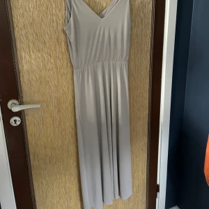 Grå långklänning - Säljer en elegant grå långklänning. Klänningen har en v-ringad hals och är ärmlös med smala axelband. Perfekt för festliga tillfällen eller en sommarkväll. Klänningen är i nyskick och har en normal passform.