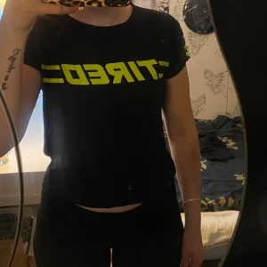 Svart T-shirt  - Svart T-shirt med neongrön text! På sidorna finns det snören. Säljer för att den inte kommer till användning