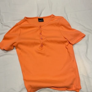 Orange ribbad topp från Gina Tricot - Säljer en superfin orange ribbad topp från Gina Tricot i storlek XS. Den har korta ärmar och knappar framtill som ger en snygg detalj. Perfekt för sommardagar eller som en färgglad bas i garderoben!