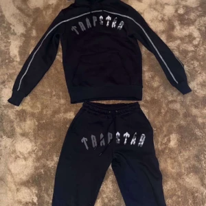 Svart mjukisbyxa och hoodie set från Trapstar - Säljer ett svart set med mjukisbyxor och hoodie från Trapstar. Båda delarna har det ikoniska Trapstar-trycket i vitt och grått. Byxorna har snörning i midjan för en justerbar passform. Perfekt för en avslappnad och stilren look!