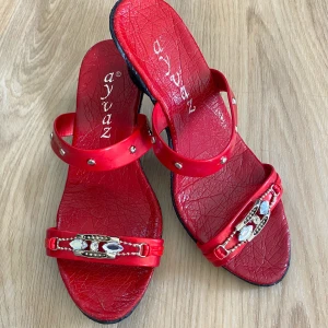 Röda sandaler med dekorativa detaljer - Säljer ett par snygga röda sandaler från Ayvaz. De har en cool design med två remmar, varav den främre är dekorerad med blingiga detaljer. Sandalerna har en svart sula med mönster för bra grepp. Perfekta för sommaren och att glida runt i på stan!
