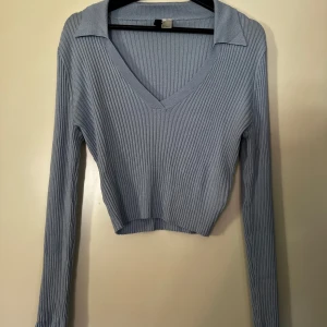 Ljusblå ribbad tröja från H&M - Säljer en superfin ljusblå ribbad tröja från H&M. Den har en snygg v-ringning och långa ärmar, perfekt för både vår och höst. Tröjan är croppad och har en stilren krage som ger en extra touch. Passar perfekt till både jeans och kjol!