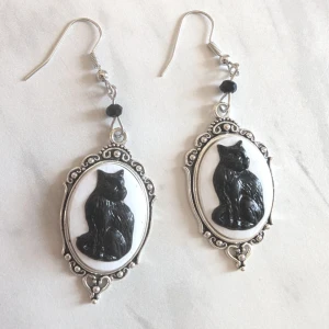 Vintage örhängen med kattmotiv - Superfina vintage örhängen med en svart katt på vit bakgrund. De har en elegant silverram med detaljer och en liten svart pärla ovanför.NY