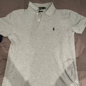Grå pikétröja från Polo - Säljer en stilren grå pikétröja från Polo i storlek M. Tröjan har korta ärmar och en klassisk krage med knappar. Perfekt för både vardag och lite finare tillfällen. Den är i bra skick och redo för en ny ägare!
