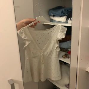 Vit blus med volangdetaljer - Säljer en söt vit blus med volangdetaljer och knyt framtill. Blusen har en v-ringad design och är ärmlös, perfekt för sommardagar. Den har ett fint broderat mönster som ger en romantisk känsla. Passar både till vardags och festliga tillfällen! Då den är lite smutsig där bak vid v ringen men bara lite!❤️