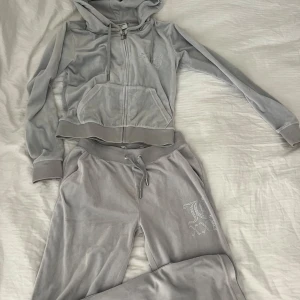 Helt ny Grå velour hoodie från Juicy Couture o byxor - Oanvänd/ny  grå velour hoodie o tillhörande byxor från Juicy Couture. Den har en dragkedja framtill och är dekorerad med glittriga stenar som bildar texten 'Juicy' på ryggen. Perfekt för en chill dag eller en avslappnad utflykt. P