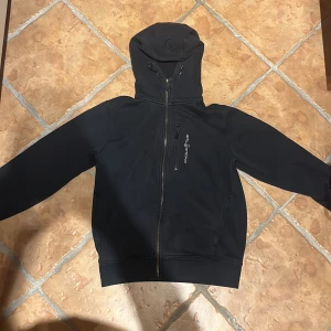 Svart hoodie från Sail Racing - Säljer en svart kofta från Sail Racing. Den har en dragkedja framtill och en praktisk huva. Perfekt för höst och vår, med en stilren design och bekväm passform. Passar både till vardags och lättare aktiviteter. Säljer den då den är för liten. Den är i storlek S, bra skick inget fel på den eller nåt. Nypris på den är 1500kr, jag säljer den för 600kr  