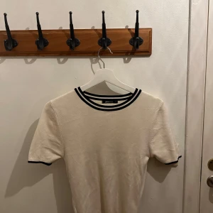 Vit kortärmad topp med svarta detaljer - Säljer en stilren vit topp från Gina Tricot med svarta ränder vid halsringningen och ärmsluten. Perfekt för både vardag och fest! Toppen är i bra skick och har en normal passform. Passar perfekt till jeans eller kjol för en avslappnad look.