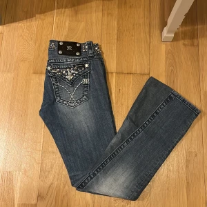 Blå jeans med nitar från Miss Me - Snygga blå jeans från Miss Me med coola nitar på fickorna och bältet. De har en lågmidjad passform och är omsydda från skinny jeans till bootcut (så var medveten om att de finns en extra söm på benen). Innebenslängden är ca 82 cm och i midjan sitter de bra på mig som är 85 cm runt midjan