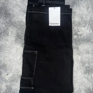 Svarta cargobyxor från Bershka - Säljer ett par svarta cargobyxor från Bershka i nytt skick. Byxorna har en loose passform med flera praktiska fickor och kontrasterande vita sömmar. Perfekta för en avslappnad och trendig stil. Passar bra till både vardag och fest!