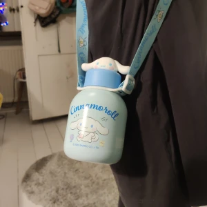 Blå Cinnamoroll flaska med axelrem - Söt och praktisk blå flaska med Cinnamoroll-motiv från Sanrio. Flaskan har en matchande axelrem för enkel bärning. Perfekt för att ta med drycker på språng. Den har ett lock med ett gulligt Cinnamoroll-ansikte. Perfekt för fans av Cinnamoroll och Sanrio!