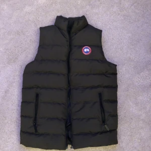  dunväst från Canada Goose - Säljer en  dunväst från Canada Goose i mycket bra skick. Västen har en dragkedja framtill och två praktiska fickor med dragkedjor. Perfekt för höst och vinter, håller dig varm och stilren. Passar både till vardags och utomhusaktiviteter.