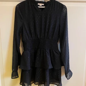 Svart klänning med volangdetaljer - Snygg svart klänning med små glittriga prickar och volangdetaljer. Den har långa ärmar och en smickrande passform som passar perfekt för höst och vinter. Klänningen är i mycket bra skick och är perfekt för både fest och vardag.