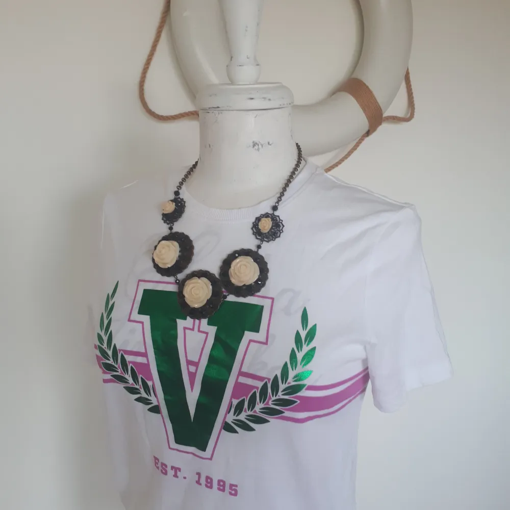 Säljer en vit t-shirt med ett stort grönt V-tryck och rosa detaljer. Texten 'EST. 1995' finns tryckt i rosa nedanför. Perfekt för en avslappnad stil eller sommarhäng. T-shirten är kortärmad och i bomull, vilket gör den skön och luftig att bära. Kolla in min profil.. T-shirts.