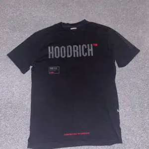 Säljer en svart t-shirt från Hoodrich i storlek S. Den har tryck både fram och bak med texten 'HOODRICH' och detaljer i rött och grått. Perfekt för en casual look eller streetwear-stil. T-shirten är i bra skick och redo för nya äventyr.