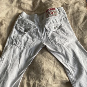 Vita jeans från True Religion - Säljer ett par vita jeans från True Religion i bra skick. De har en klassisk femficksdesign med snygga detaljer på bakfickorna. Jeansen är tillverkade i USA och har en normal passform. Perfekta för både vardag och fest. Saknar knapp vilket lätt kan sys på. 