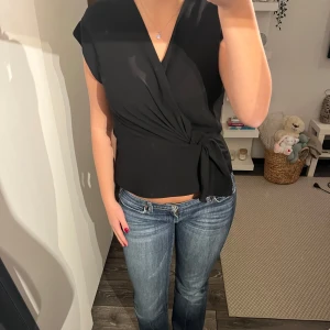 Svart omlottopp från Zara - Snygg svart omlottopp från Zara i storlek S. Toppen har en elegant v-ringning och knytning i midjan som ger en jättefin passform. Perfekt för både vardag och fest!