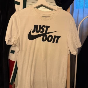 Vit t-shirt från Nike - Säljer en klassisk vit t-shirt från Nike med det ikoniska 'Just Do It'-trycket i svart på framsidan. T-shirten är i bomull och har en normal passform med korta ärmar. Perfekt för både träning och vardag. 🏃‍♂️