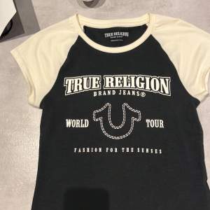 Säljer en snygg svart och vit t-shirt från True Religion i storlek XS. Den har ett coolt tryck med texten 'World Tour' och 'Fashion for the Senses'. Perfekt för en avslappnad stil. T-shirten är kortärmad och i mycket bra skick.