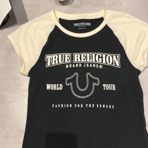 Svart och vit t-shirt från True Religion - Säljer en snygg svart och vit t-shirt från True Religion i storlek XS. Den har ett coolt tryck med texten 'World Tour' och 'Fashion for the Senses'. Perfekt för en avslappnad stil. T-shirten är kortärmad och i mycket bra skick.