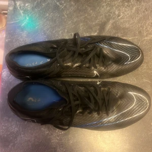 Svarta Nike Mercurial fotbollsskor - Säljer ett par svarta Nike Mercurial fotbollsskor i mycket bra skick. Skorna har en snygg och modern design med snörning och en slitstark sula för bästa grepp på planen. Perfekta för fotbollsspelare som vill ha både stil och funktion.  