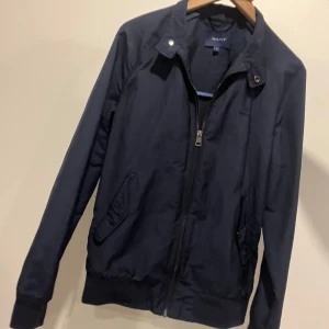 Mörkblå jacka från GANT - Säljer en stilren mörkblå jacka från GANT i mycket bra skick. Jackan har en dragkedja framtill och knappar vid kragen. Perfekt för höst och vår med sin klassiska design och bekväma passform.