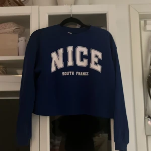 Blå croppad tröja från H&M - Säljer en snygg blå croppad tröja från H&M med texten 'NICE SOUTH FRANCE' på framsidan. Perfekt för en avslappnad stil och passar bra till jeans eller kjol. Tröjan har långa ärmar och är i bra skick. Perfekt för vår och sommar!