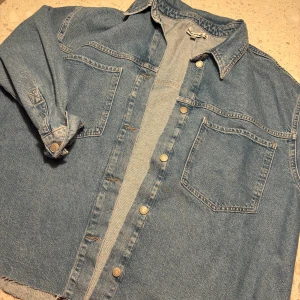 Jeansskjorta från Denim Rebel - Säljer en snygg jeansskjorta från Denim Rebel i storlek XL. Skjortan har en klassisk blå denimfärg och är utrustad med knappar framtill. Perfekt för en avslappnad stil eller som en lätt jacka under vår och höst.