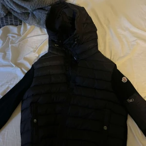 Svart cardigan från Moncler - Säljer en snygg svart cardigan från Moncler i storlek L. Jackan har en quiltad design med dragkedja och en huva för extra värme. Perfekt för höst och vinter! Den är i mycket bra skick och redo för nya äventyr. 