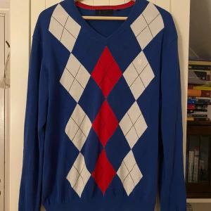Blå tröja med argylemönster från Riley - Säljer en snygg blå tröja från Riley med klassiskt argylemönster i rött och vitt. Tröjan har en v-ringad hals och långa ärmar, perfekt för både höst och vinter. Den är i mycket bra skick och passar perfekt för en stilren look😎