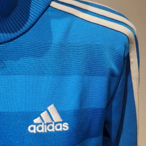 Blå randig jacka från Adidas - Säljer en snygg blå randig jacka från Adidas med klassiska vita ränder på ärmarna. Jackan har en dragkedja framtill och är långärmad med ribbade muddar. Perfekt för träning eller som en stilren vardagsjacka. Passar bra till både sport och casual outfits!