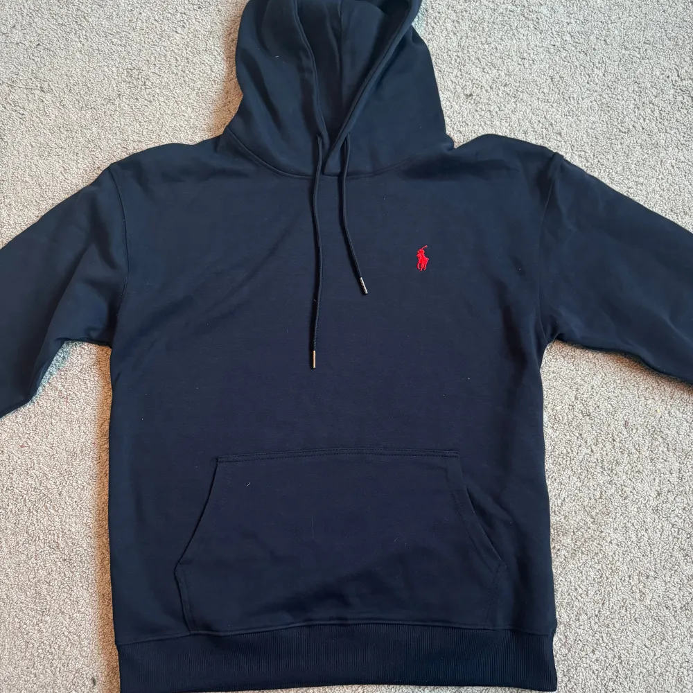 Säljer en snygg svart hoodie från Ralph Lauren i mycket bra skick. Den har en klassisk design med en röd broderad logga på bröstet och en praktisk magficka. Perfekt för både vardag och chill. Passar till allt och är superbekväm!. Tröjor & Koftor.