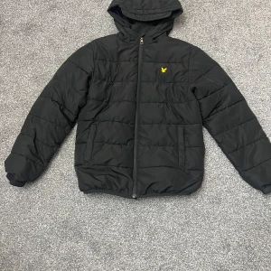 Svart pufferjacka med huva - Säljer en svart pufferjacka från Lyle & Scott i mycket bra skick. Jackan har en praktisk huva och dragkedja framtill. Perfekt för kyliga dagar och håller dig varm och bekväm. Den har en gul logga på bröstet som ger en snygg detalj.