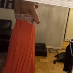 Orange axelbandslös klänning från Jovani - Säljer en orange/rosa klänning från Jovani. Den har en glittrig överdel med paljetter och en lång, flödande kjol. Perfekt för festliga tillfällen som bal dop bröllop m.m. Använd endast en gång. Den är alldeles för stor för mig så ser inte jätte bra ut nu på bilden😁