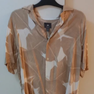 Beige och vit mönstrad skjorta från H&M - Säljer en snygg beige och vit skjorta från H&M med ett abstrakt mönster. Skjortan har korta ärmar och knappar framtill. Perfekt för sommardagar eller en avslappnad fest. Materialet känns lätt och luftigt, vilket gör den idealisk för varmare väder. Passar bra till både jeans och shorts!