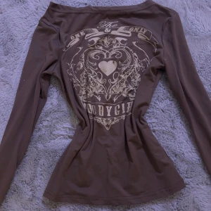 Brun långärmad topp med tryck - Säljer en brun långärmad topp med ett coolt tryck på ryggen. Perfekt för höst och vår, den har en normal passform och är superbekväm. Trycket har ett hjärta och text som ger en edgy look. Passar både till vardags och fest!