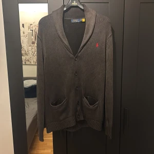  Ralph Lauren cardigan  - Säljer en snygg grå cardigan från Polo Ralph Lauren i mycket bra skick. Den har en klassisk design med knappar och två fickor framtill. Den röda loggan på bröstet ger en stilren touch. Perfekt för höst och vinter!                                           Ställ gärna frågor om det undrad!!! 