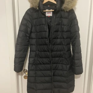 Svart dunjacka från Tommy Jeans - Säljer en svart lång dunjacka från Tommy Jeans i storlek M. Jackan har en mysig pälskantad huva och dragkedja framtill. Perfekt för kalla vinterdagar! 🖤