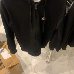 Säljer en svart hoodie från Moncler i mycket bra skick. Den har en dragkedja framtill och en liten Moncler-logga på bröstet. Perfekt för höst och vår, med en stilren och enkel design. Passar både till vardags och lite mer avslappnade tillfällen. Skick10/10