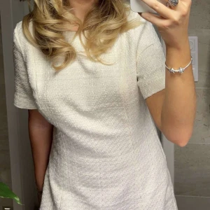 Vit klänning i texturerat tyg - Säljer en elegant vit klänning i texturerat tyg. Den har en klassisk och stilren design med korta ärmar och en figurnära passform. Perfekt för både vardag och festliga tillfällen. Klänningen är i mycket bra skick! Stl 38. Aldrig använd och prislappen är kvar 