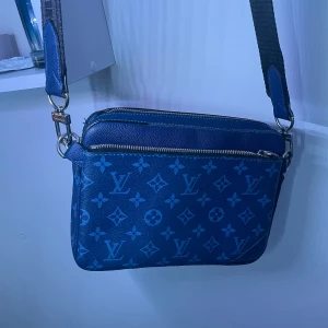 Blå axelväska med monogram - Snygg blå axelväska med klassiskt monogrammönster. Väskan har en justerbar axelrem och en praktisk dragkedja upptill. Perfekt för både vardag och festliga tillfällen. Den är i mycket bra skick och redo att användas direkt!