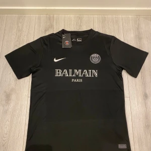 PSG x Balmain fotbollströja - Säljer en svart PSG x Balmain fotbollströja i Dri-FIT material. Tröjan har korta ärmar och är i nytt skick med etiketter kvar. Perfekt för fotbollsfans som vill ha något unikt och stilrent. Stor logga på bröstet och snygg design. Passar både till match och vardag! Tröjan är sällsynt och går ej att köpa i butik. 