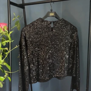 Svart glittrig topp - Snygg svart glittrig topp med långa ärmar, perfekt för festliga tillfällen. Toppen har en croppad passform och en liten öppning i ryggen som ger en extra detalj. Den är både stilren och iögonfallande, idealisk för en utekväll eller nyårsfest.