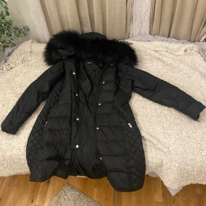 Svart dunjacka från Rockandblue - Säljer en snygg svart dunjacka från Rockandblue i storlek 44. Jackan har en mysig pälskantad huva och quiltad design. Den är perfekt för kalla vinterdagar och har både dragkedja och knappar för extra värme. Ett måste för vintern! ❄️