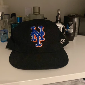Svart keps från New Era - Säljer en svart New Era keps med broderad logga i blått och orange på framsidan. Kepsen är i bra skick och har MLB-loggan på baksidan. Perfekt för sommarens alla äventyr eller för att visa ditt stöd för ditt favoritlag!