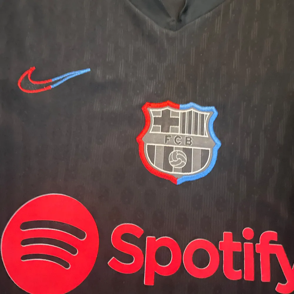 Säljer en svart FC Barcelona tröja med korta ärmar. Den har röda och blå detaljer på sidorna och ett tryck med 'Lewandowski 9' på ryggen. Framtill finns Spotify-loggan och klubbmärket. Tröjan är i Dri-FIT material, perfekt för träning eller match!. T-shirts.